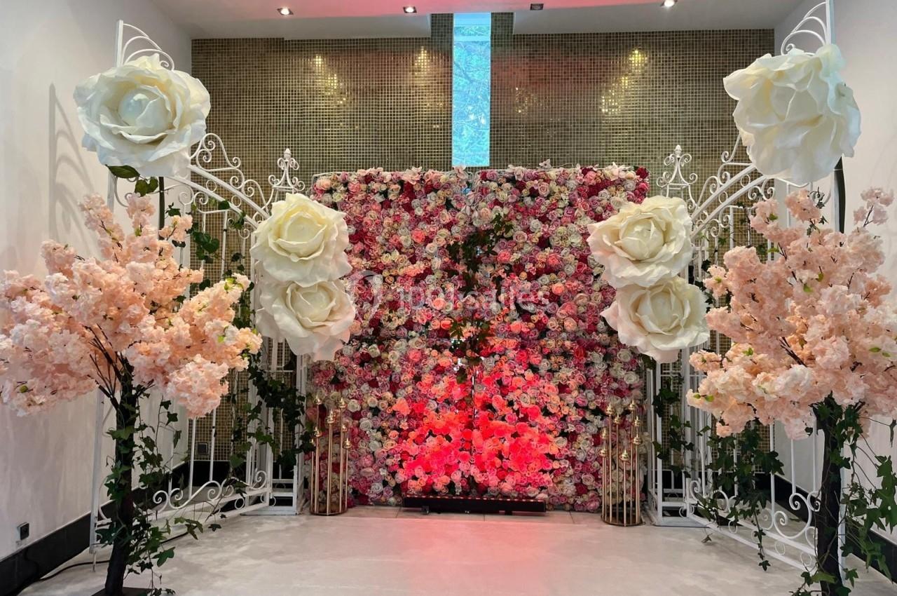 Mur floral rose et blanc encadré de structures métalliques décoratives, avec des arbres artificiels fleuris.