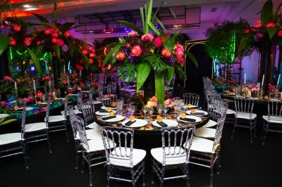 Table dressée avec des chaises transparentes, ornée de compositions florales tropicales colorées et de bougies.