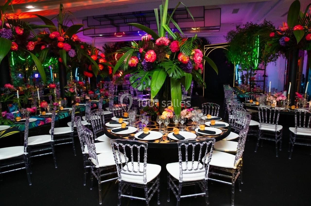Salle de réception décorée avec des tables rondes, des chaises transparentes et des arrangements floraux colorés.