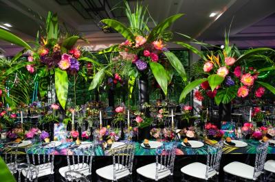 Table dressée avec des chaises transparentes, ornée de compositions florales tropicales colorées et de bougies.