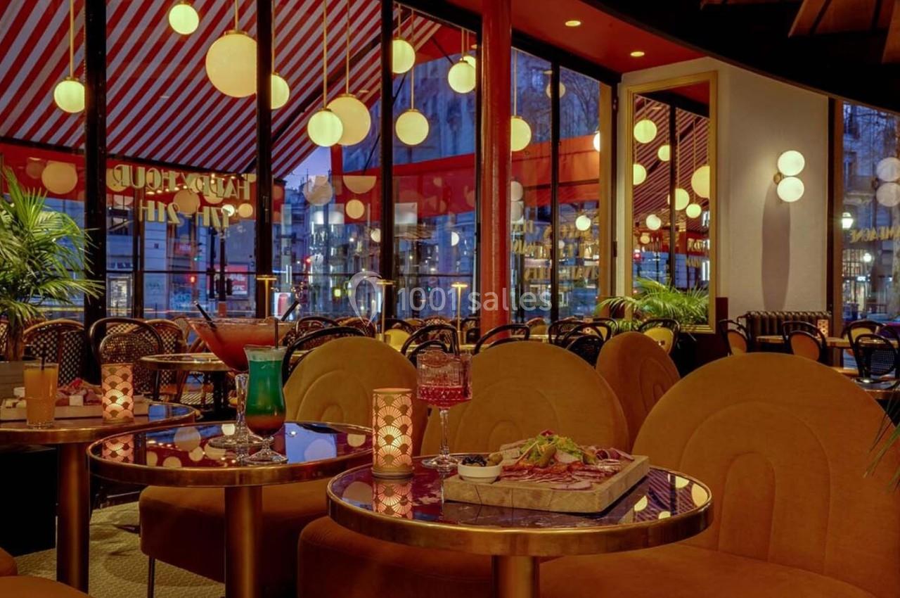 Salle de restaurant avec des tables dorées, fauteuils orange, éclairage chaleureux et grandes baies vitrées donnant sur l…