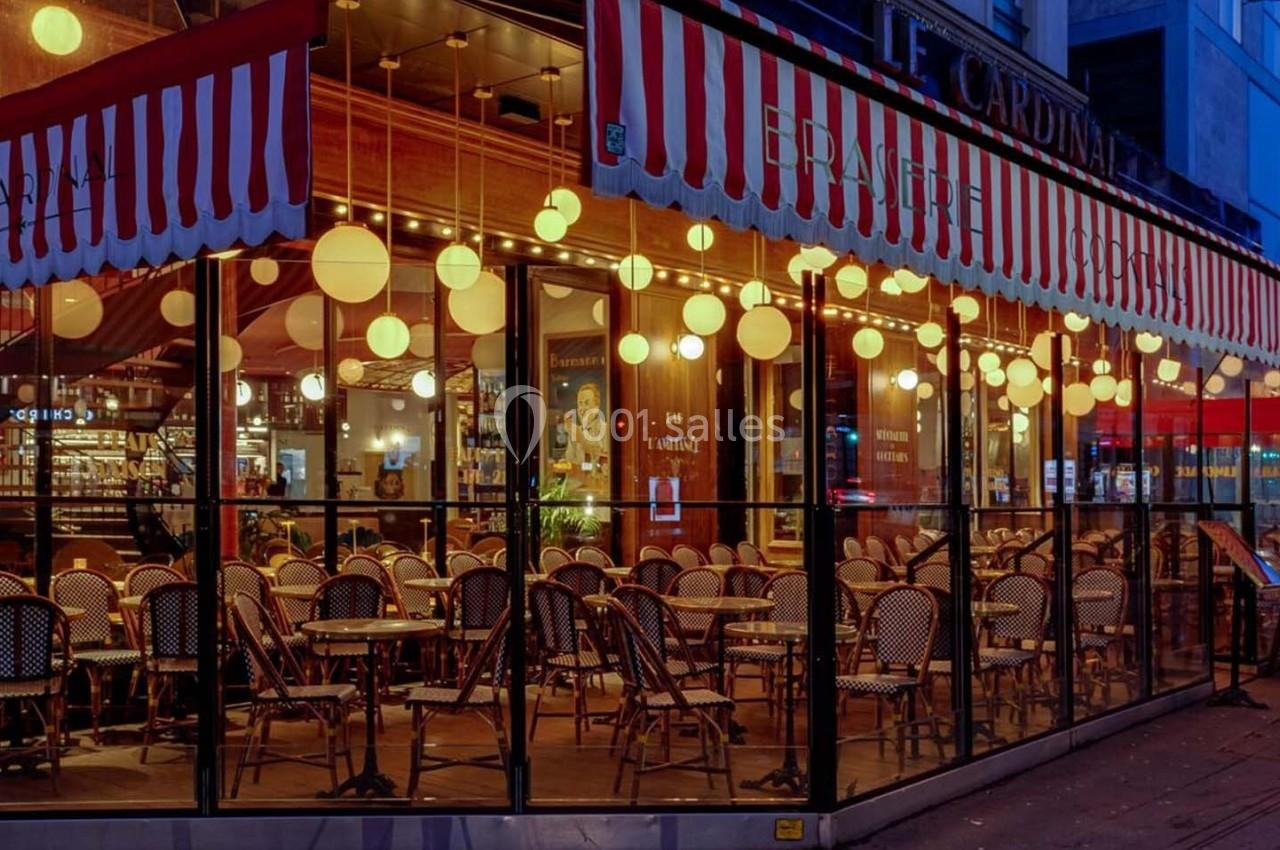 Terrasse d'un café-restaurant avec chaises en osier et lampes suspendues, vue à travers des baies vitrées le soir.