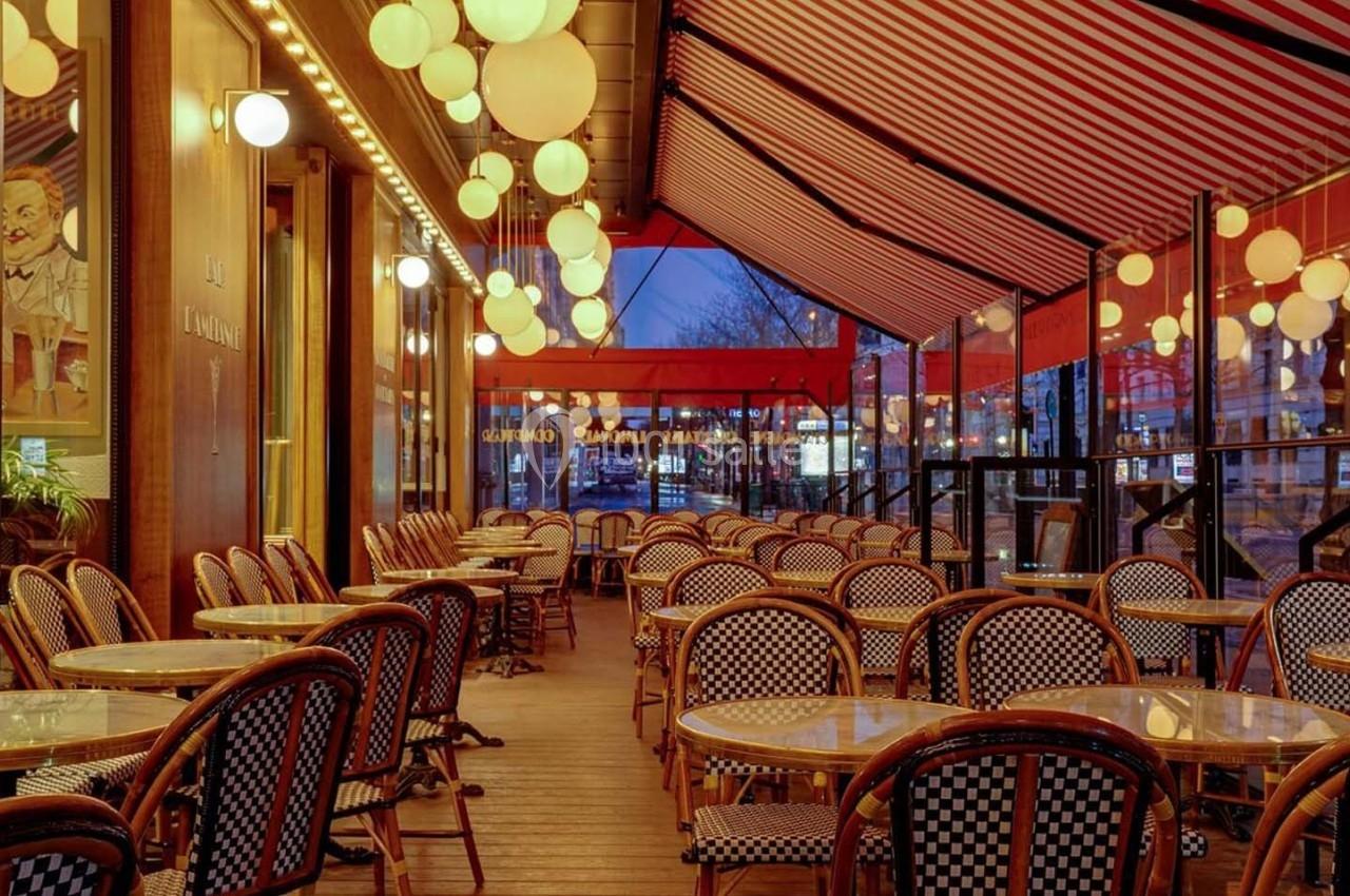 Terrasse de café avec tables et chaises en osier, éclairée par des lampes suspendues sous un auvent rayé rouge et blanc.