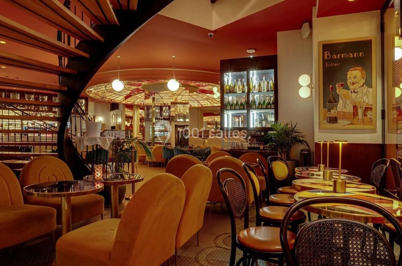 Salle de restaurant chaleureuse avec fauteuils en velours, tables rondes, escalier en colimaçon et éclairage tamisé.
