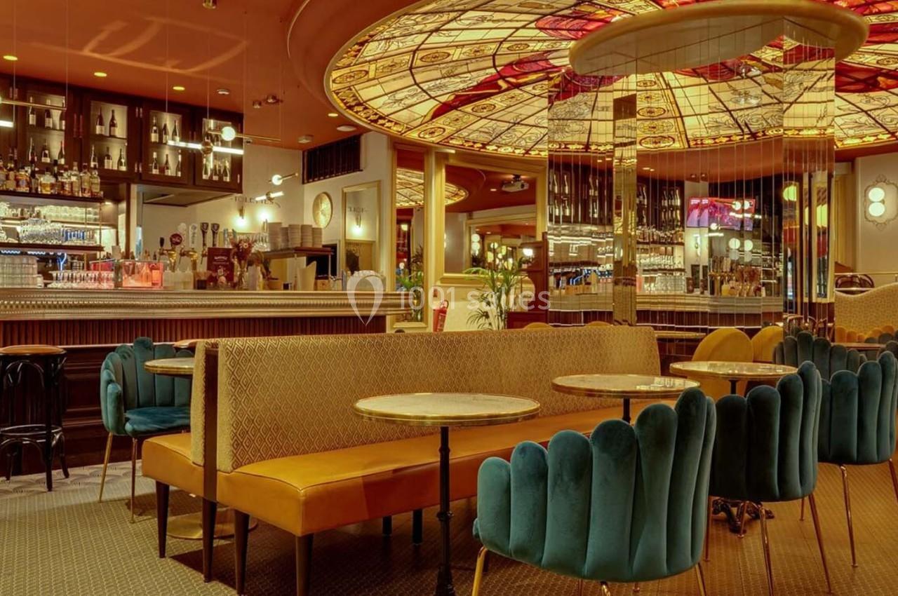 Intérieur d'un café avec banquettes, fauteuils colorés et un plafond décoré de vitraux lumineux.