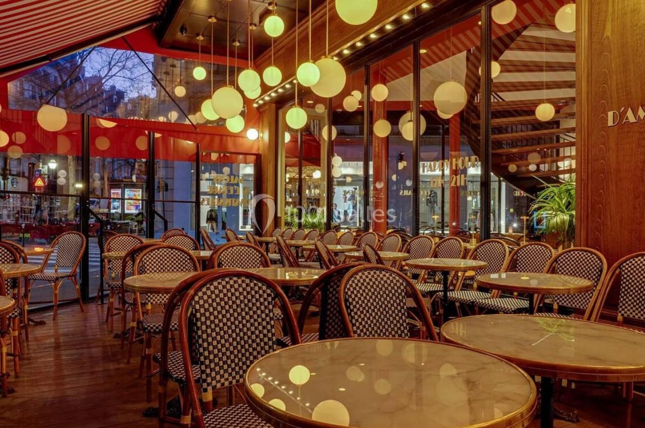 Salle de restaurant avec tables rondes, chaises en osier et luminaires suspendus, vue sur l'extérieur vitré.