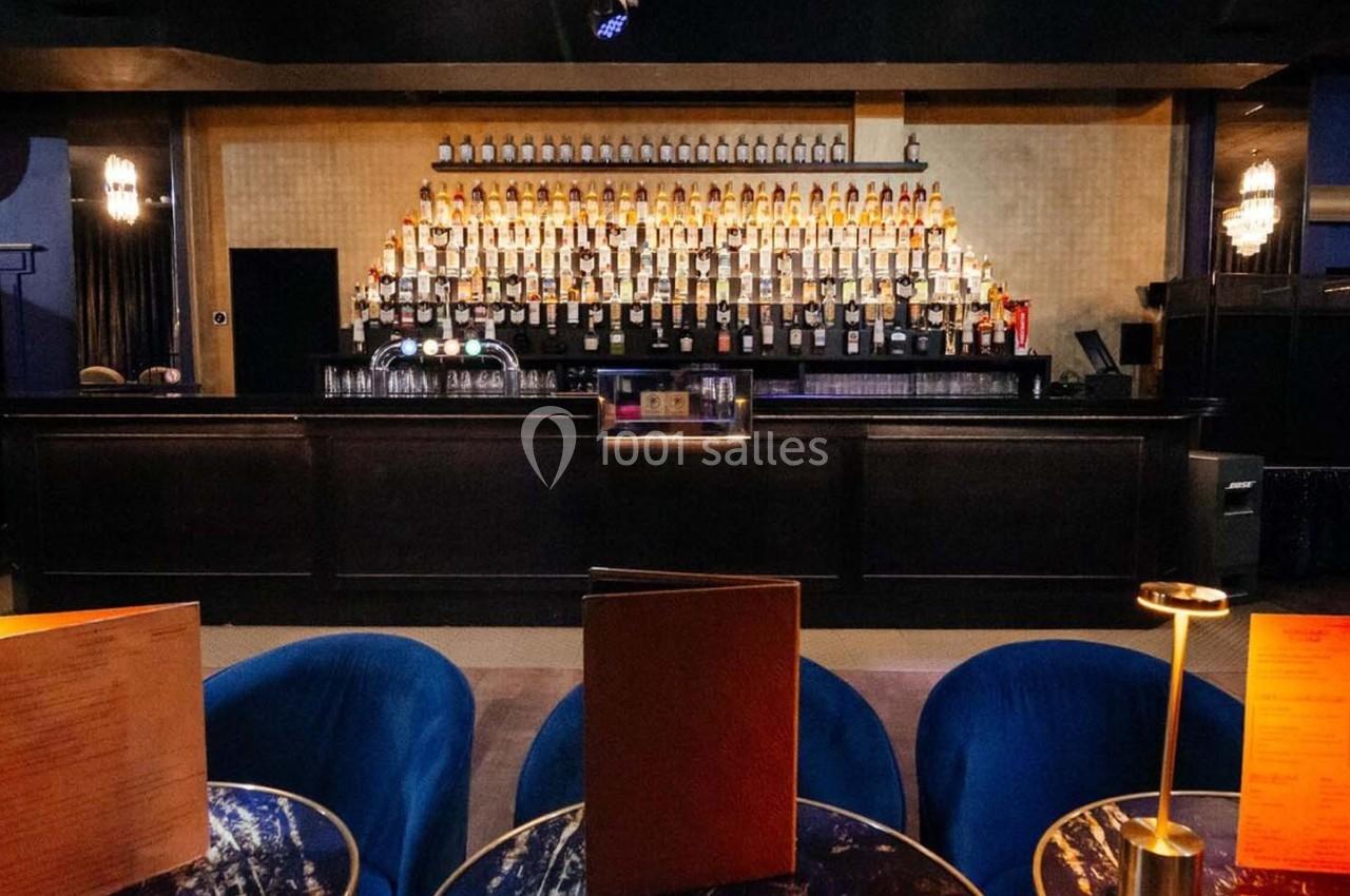 Bar élégant avec un long comptoir, des étagères remplies de bouteilles et des tables avec lampes et menus au premier plan.