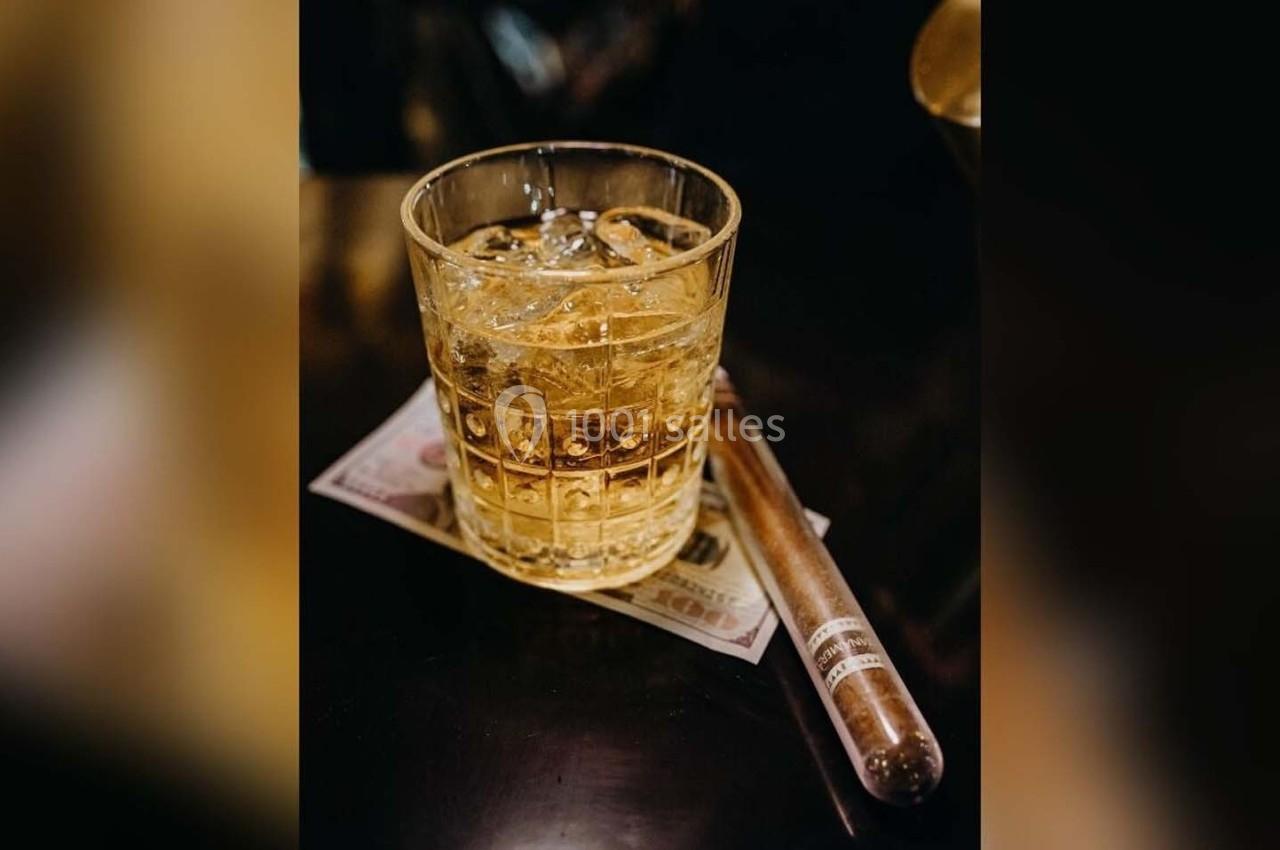 Verre de whisky avec glaçons posé sur un billet, accompagné d'un cigare sur une table sombre.