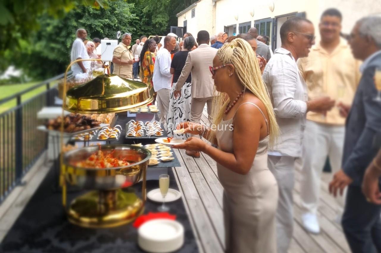Des convives discutent et se servent à un buffet en extérieur, avec des plats variés et une ambiance conviviale.
