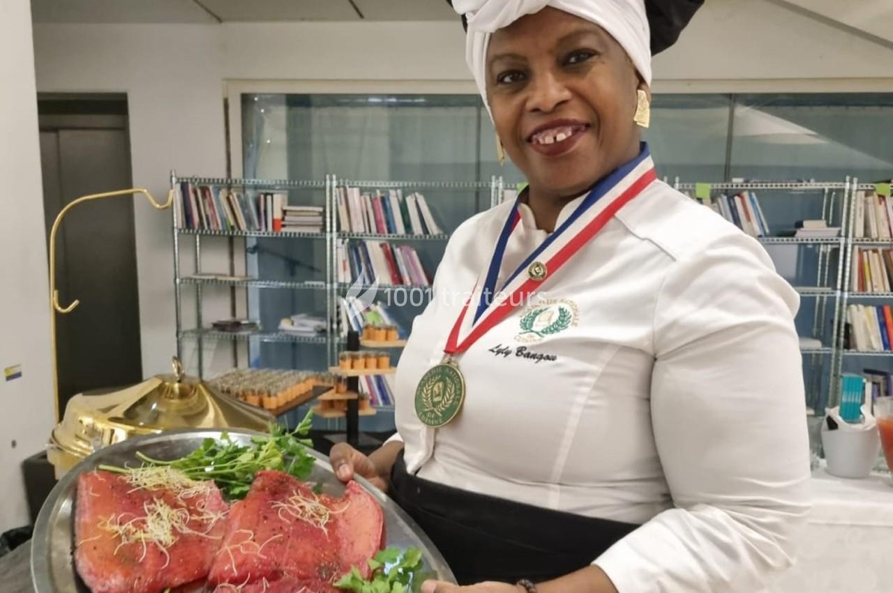 Une cheffe en tenue professionnelle présente un plateau de viande garnie de pousses, dans une cuisine avec des étagères en…