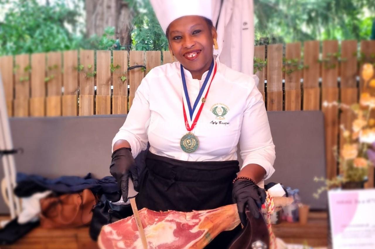 Une cheffe en tenue blanche et toque découpe un jambon sur un stand en extérieur, portant une médaille autour du cou.
