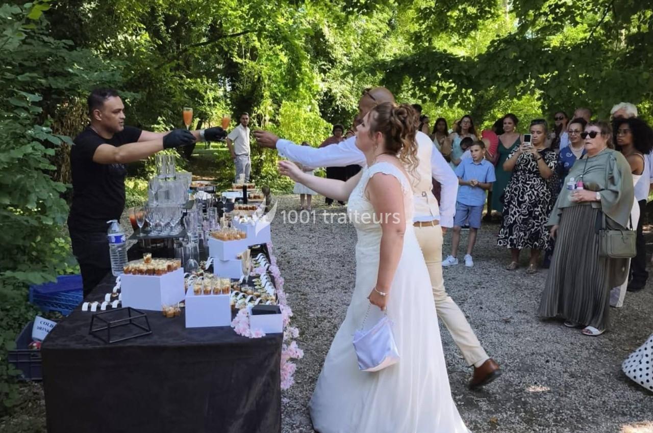 Un couple en tenue de mariage trinque avec un serveur devant une table de buffet en extérieur, entouré d'invités.