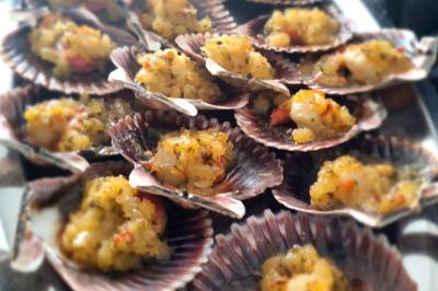 Huîtres gratinées garnies de fromage et épices, présentées sur un plateau en bois, avec légumes en arrière-plan.