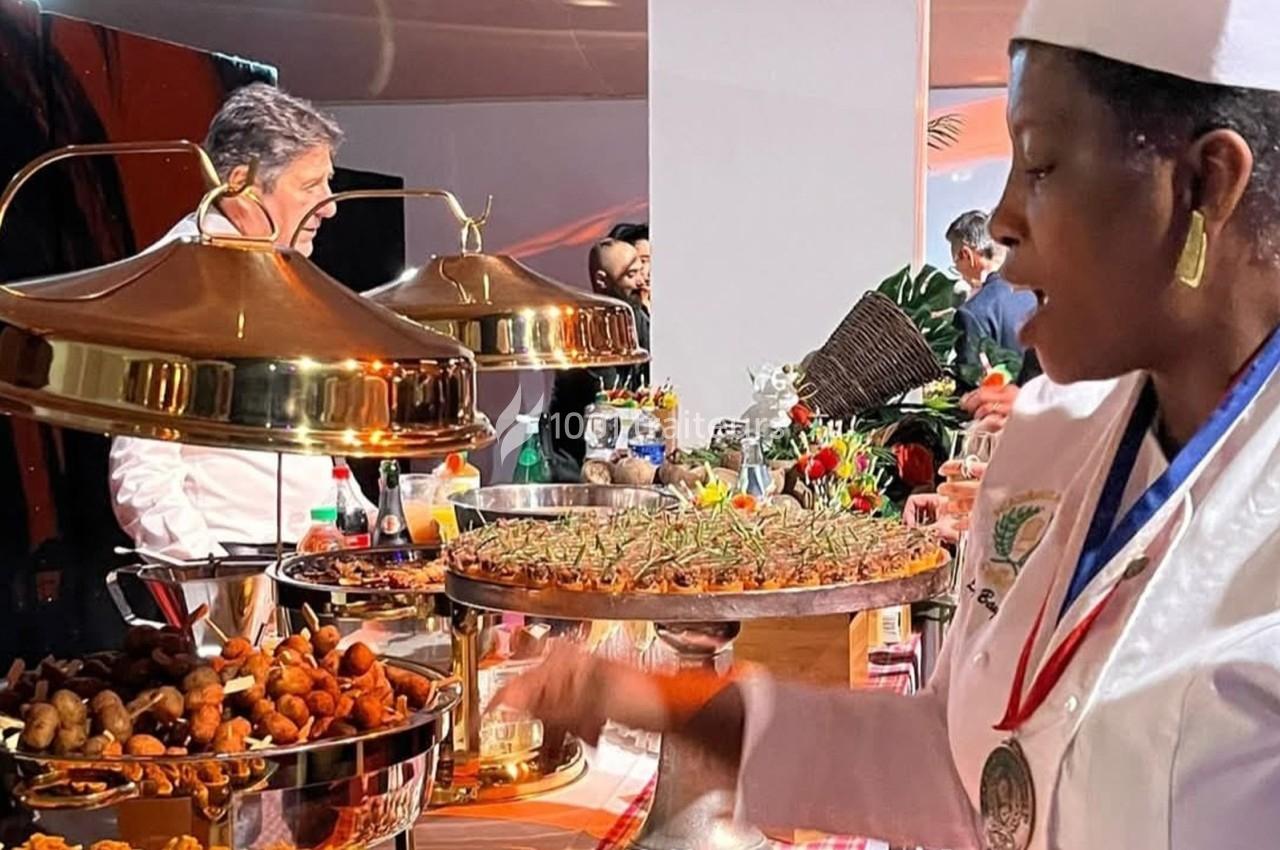 Un chef et une cuisinière en tenue professionnelle préparent et présentent des plats sur un buffet garni.