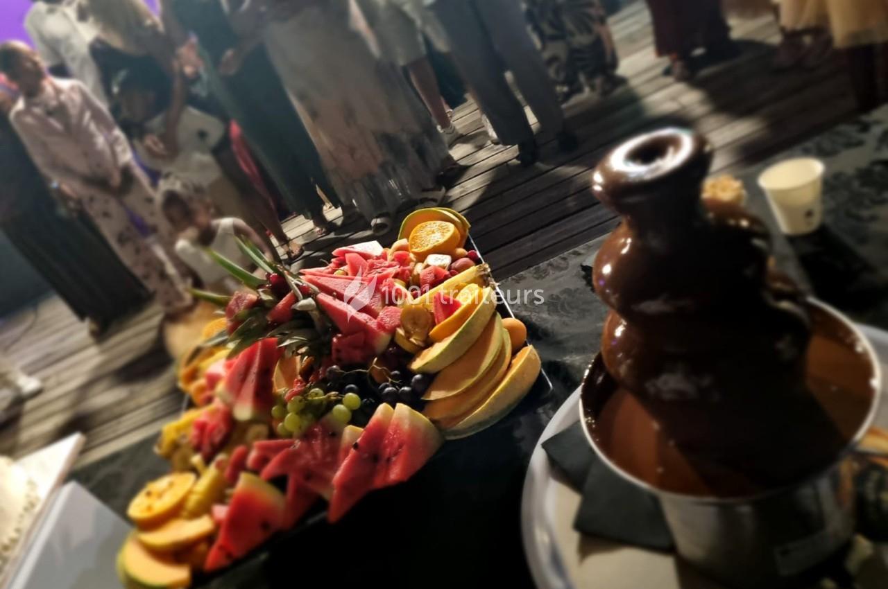Plateau de fruits variés et fontaine de chocolat au premier plan, avec des personnes en arrière-plan flou.