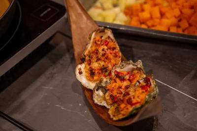 Huîtres gratinées garnies de fromage et épices, présentées sur un plateau en bois, avec légumes en arrière-plan.