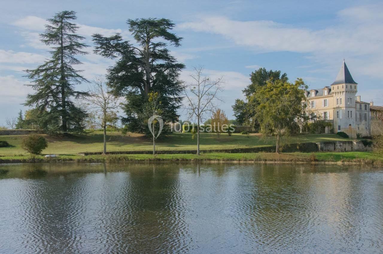 Vue d'un château entouré d'arbres, avec un étang au premier plan reflétant le paysage.