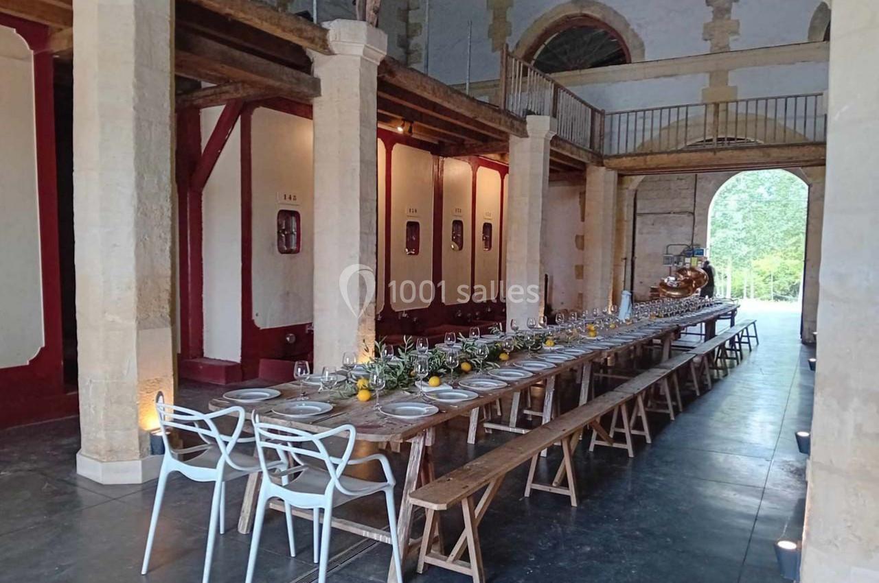 Grande table en bois dressée pour un repas dans une salle aux murs en pierre avec des cuves de vin en arrière-plan.