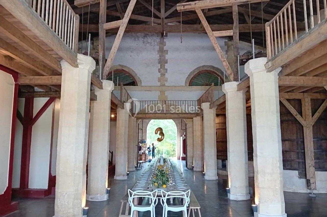 Grande salle avec charpente en bois apparente, table longue dressée au centre et décorations lumineuses.