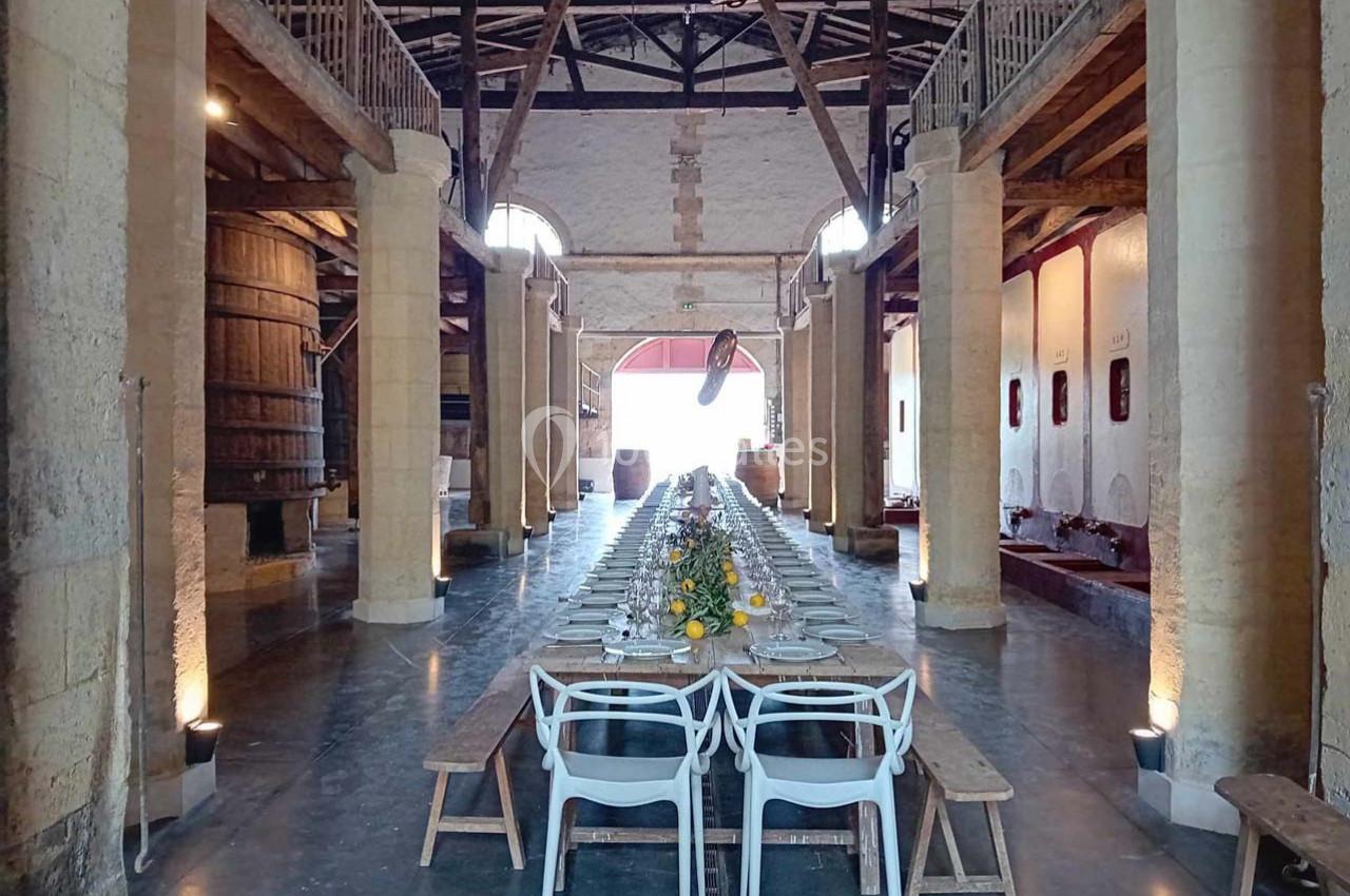 Grande salle avec une longue table dressée pour un repas, entourée de bancs et de chaises, dans un cadre rustique avec…