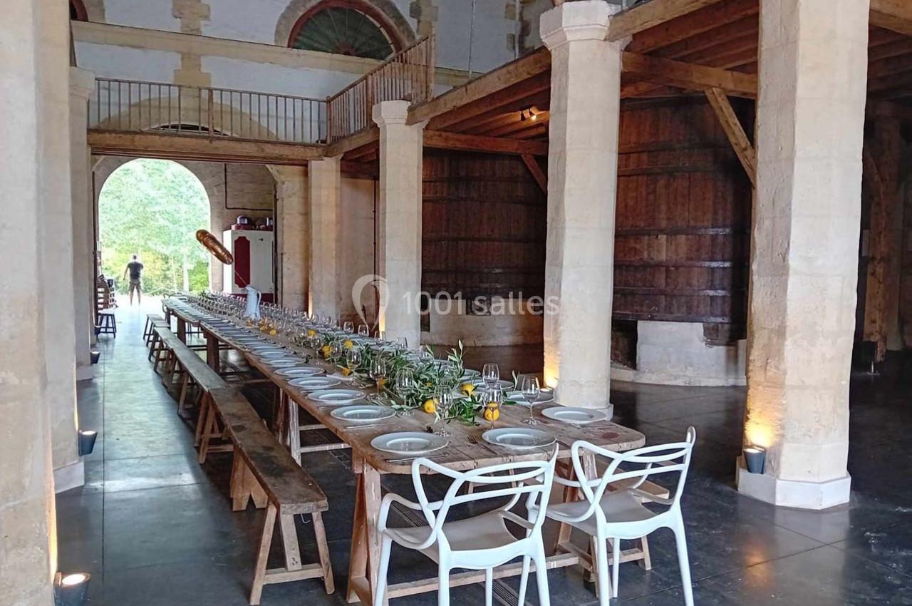 Grande table en bois dressée avec vaisselle et décorations florales dans une salle rustique avec poutres et tonneaux.