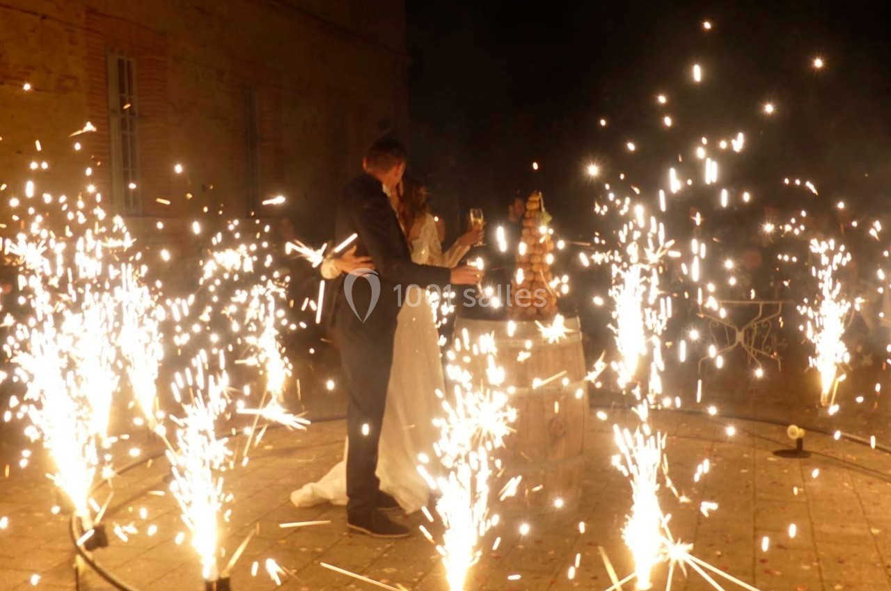 Un couple en tenue de mariage trinque devant une pièce montée, entouré de fontaines lumineuses en extérieur.