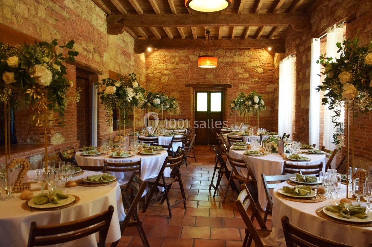 Salle de réception rustique avec tables rondes dressées, décorées de fleurs et nappes blanches, éclairée par une lumière…