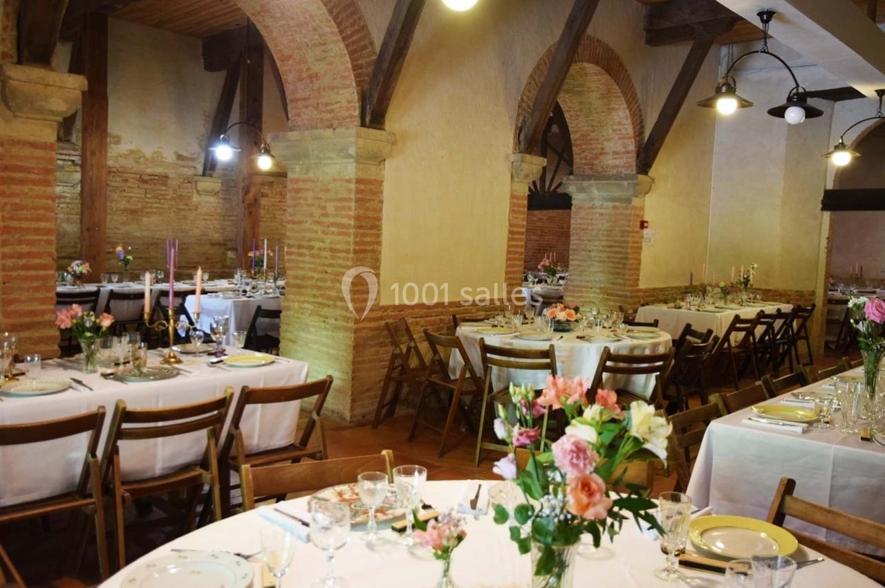 Salle de réception avec tables décorées de fleurs, nappes blanches et chaises en bois, sous des arches en briques.