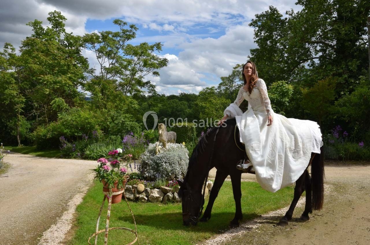 Une femme en robe blanche est assise sur un cheval noir qui broute de l'herbe, dans un cadre verdoyant et fleuri.