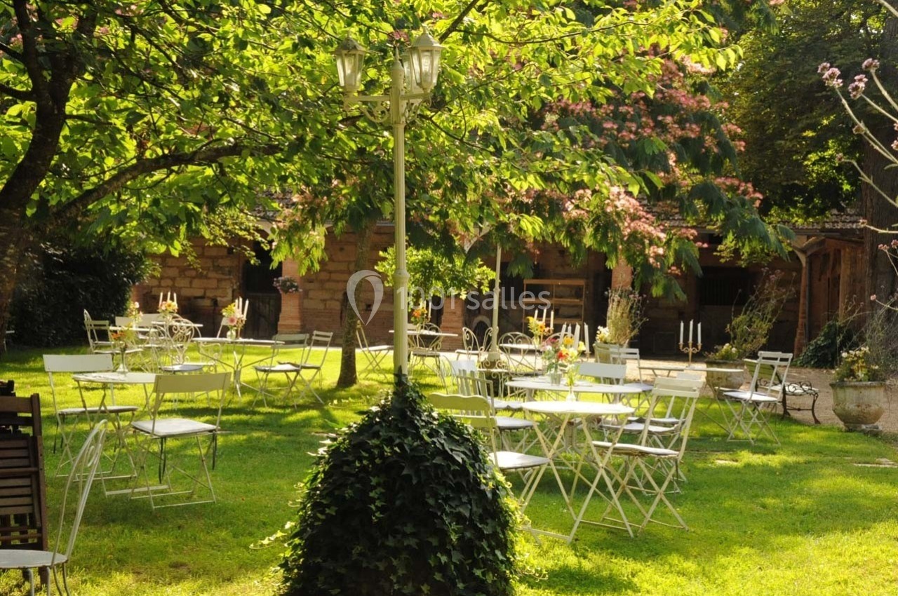 Chaises et tables blanches disposées dans un jardin verdoyant, entouré d'arbres et éclairé par un lampadaire.
