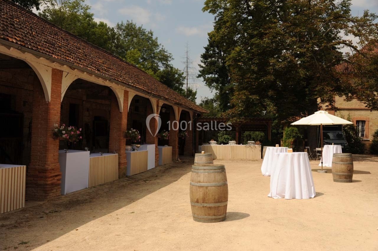 Cour extérieure avec tables hautes nappées, tonneaux en bois et buffets sous une galerie couverte, par temps ensoleillé.
