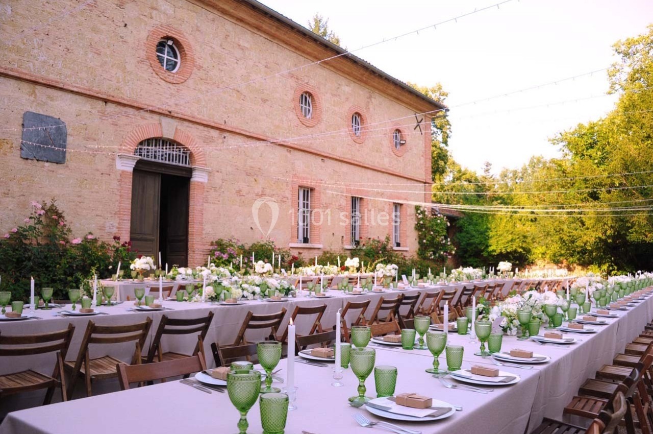 Grande table extérieure dressée avec vaisselle verte et décor floral, devant un bâtiment en pierre et un jardin verdoyant.