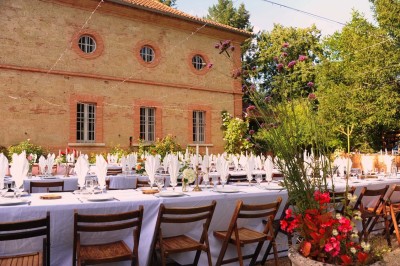 Grande table dressée avec vaisselle élégante, pains individuels et décoration florale, sous une terrasse en bois ouverte sur…