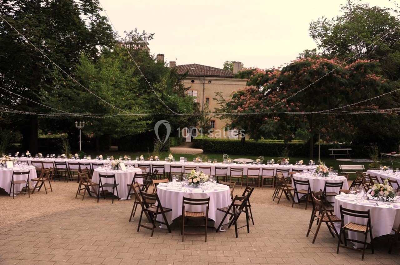 Tables rondes dressées avec nappes blanches et chaises en bois dans un jardin, décorées pour un événement en plein air.