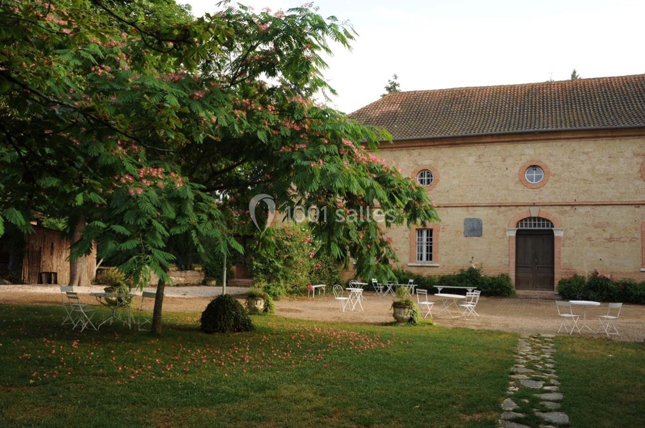 Cour avec des tables et chaises blanches sous un grand arbre, devant un bâtiment en briques avec une porte en bois.