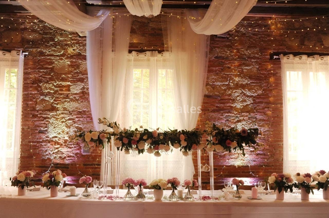 Table décorée pour un mariage avec fleurs, bougies et guirlandes lumineuses, devant un mur de briques et des rideaux blancs.