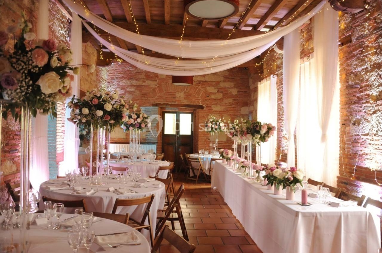 Salle de réception décorée pour un mariage avec tables dressées, fleurs et guirlandes lumineuses suspendues.