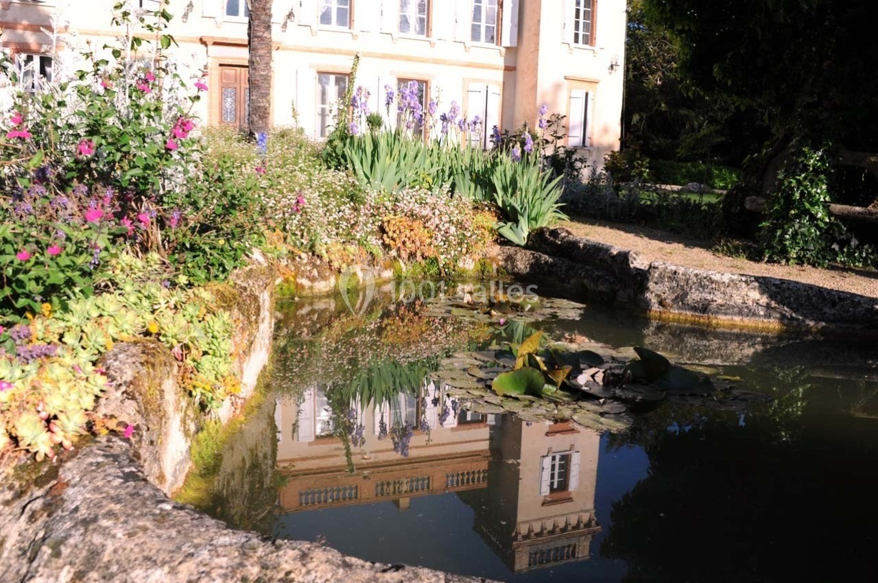 Étang entouré de fleurs et de végétation, reflétant une façade de bâtiment beige sous un ciel ensoleillé.