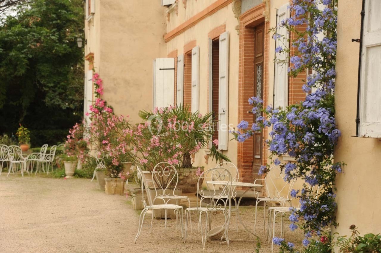 Façade d'une maison ancienne avec volets blancs, mobilier de jardin en fer forgé et plantes fleuries colorées.