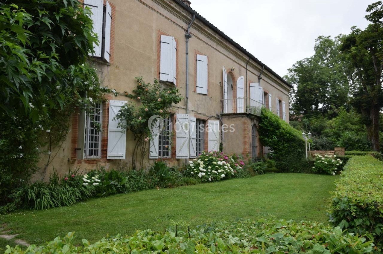 Façade d'une maison ancienne avec volets blancs, jardin verdoyant et massifs de fleurs devant.