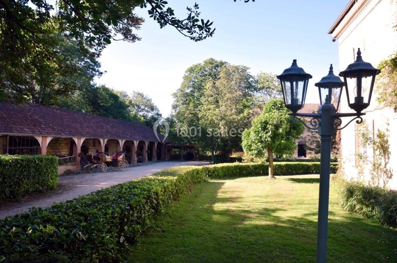 Allée bordée de bâtiments en pierre, avec une calèche stationnée et un lampadaire dans un jardin verdoyant.