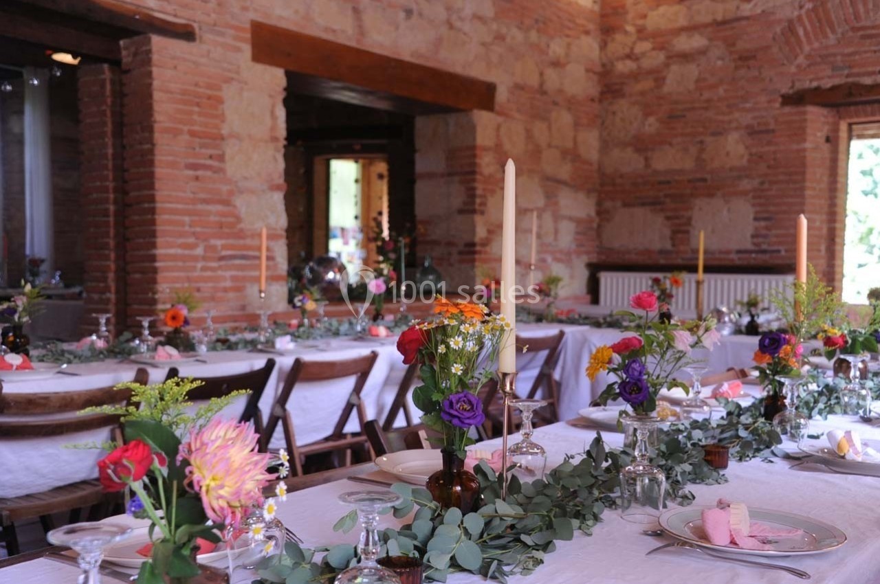 Salle en pierre aménagée pour un repas, avec tables décorées de fleurs colorées, bougies et nappes blanches.