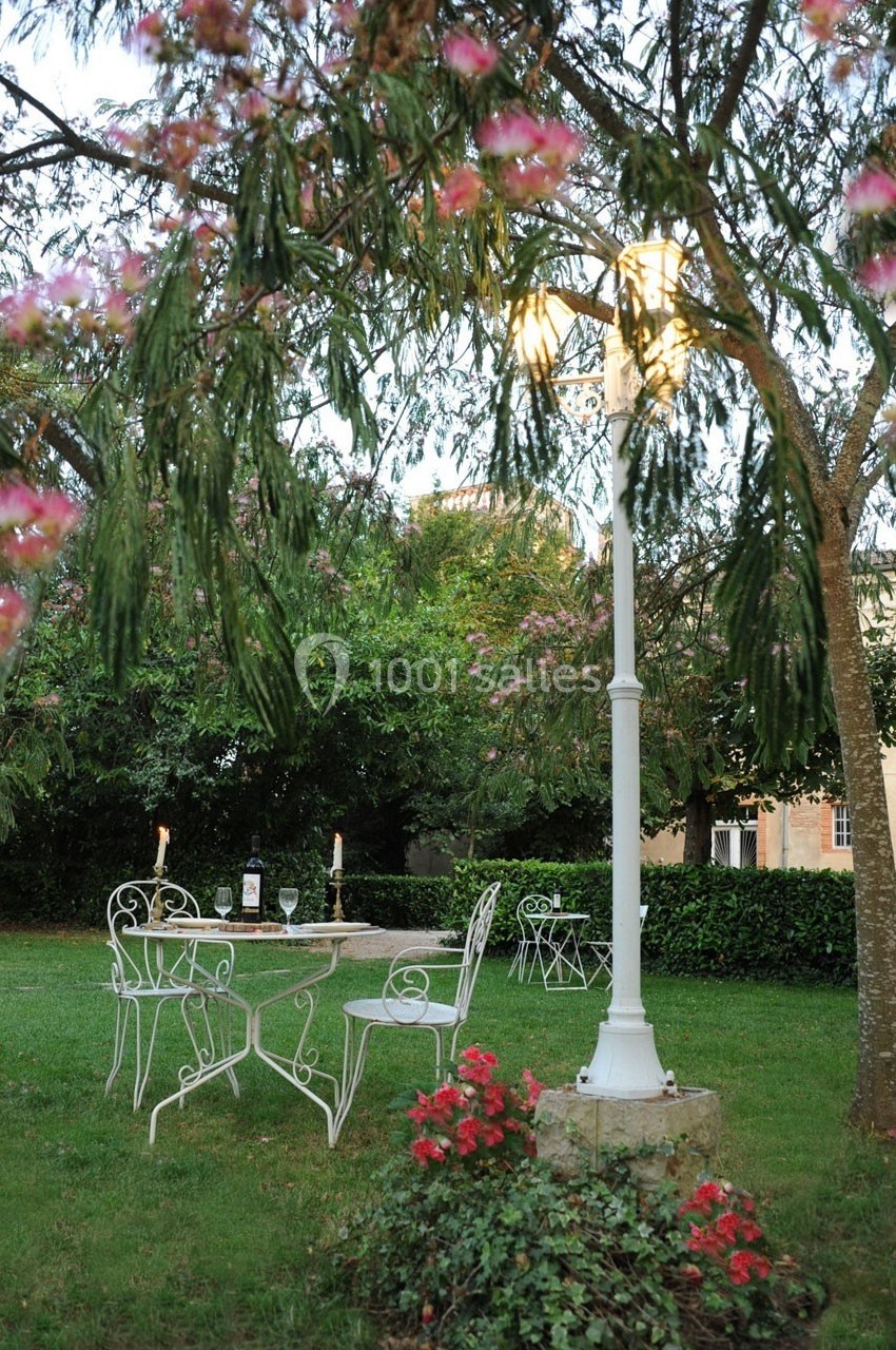 Mobilier de jardin en fer forgé blanc sous un lampadaire allumé, entouré de végétation et de fleurs rouges.