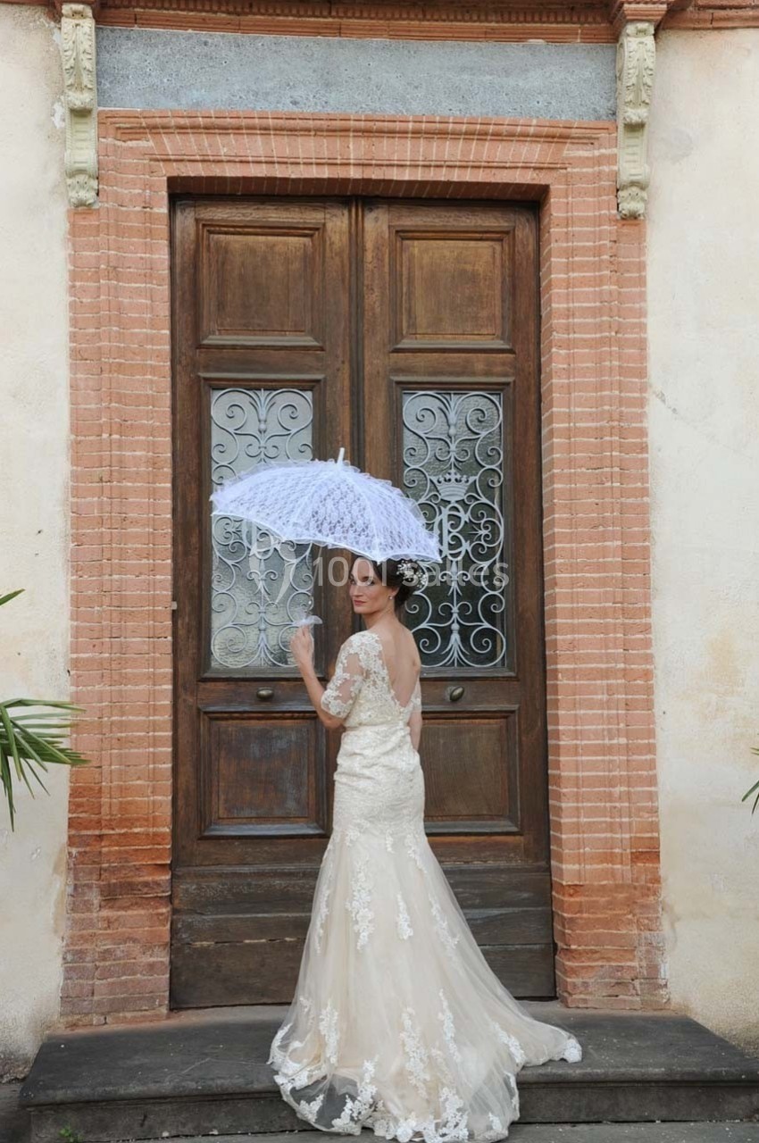 Femme en robe de mariée tenant une ombrelle blanche devant une porte en bois ornée de motifs en fer forgé.