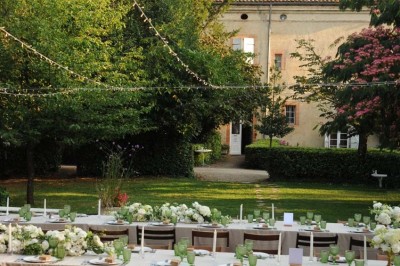 Grande table dressée avec vaisselle élégante, pains individuels et décoration florale, sous une terrasse en bois ouverte sur…