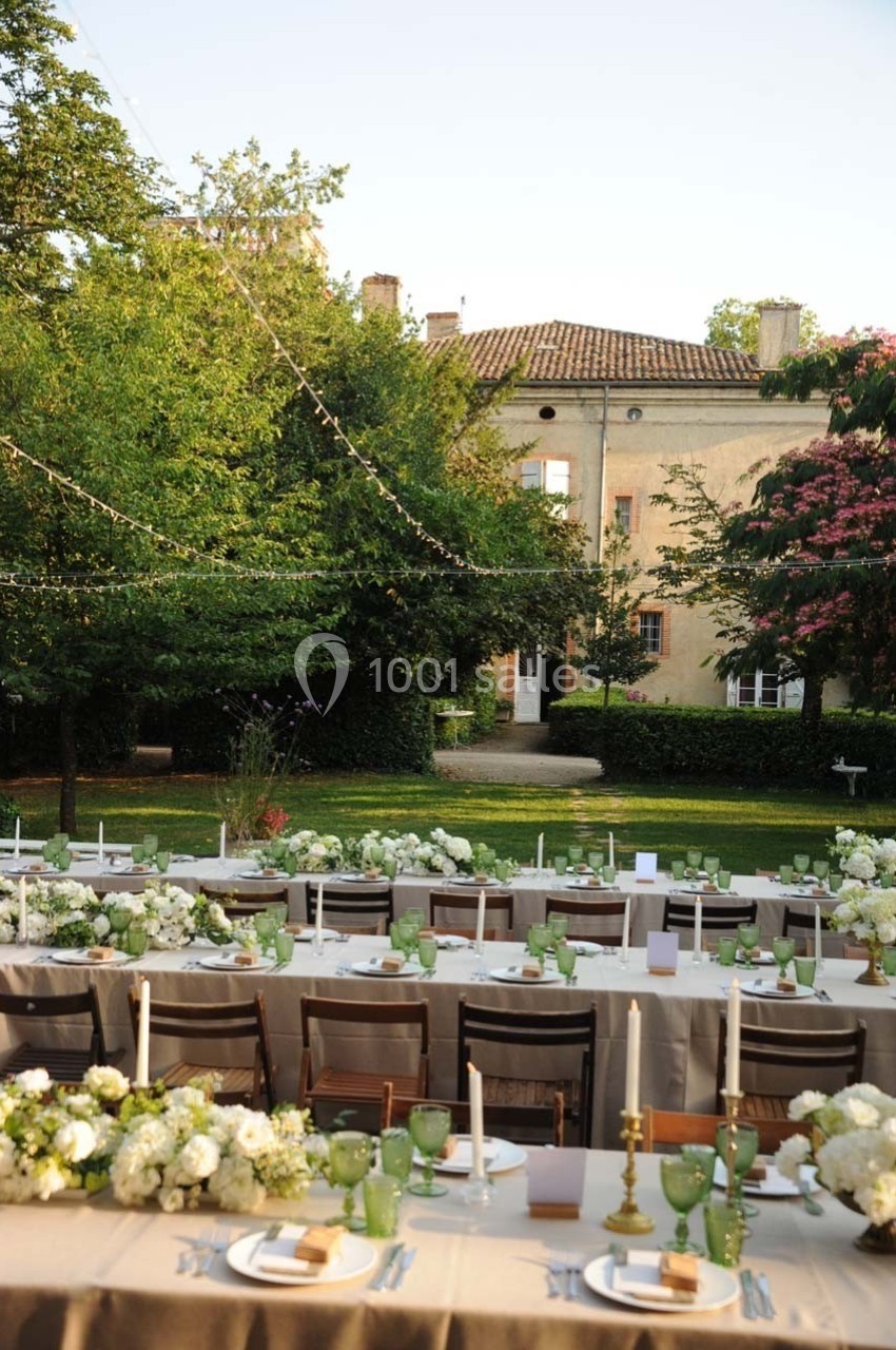 Tables décorées pour un repas en plein air avec nappes beiges, fleurs blanches et bougies, dans un jardin devant un bâtiment…