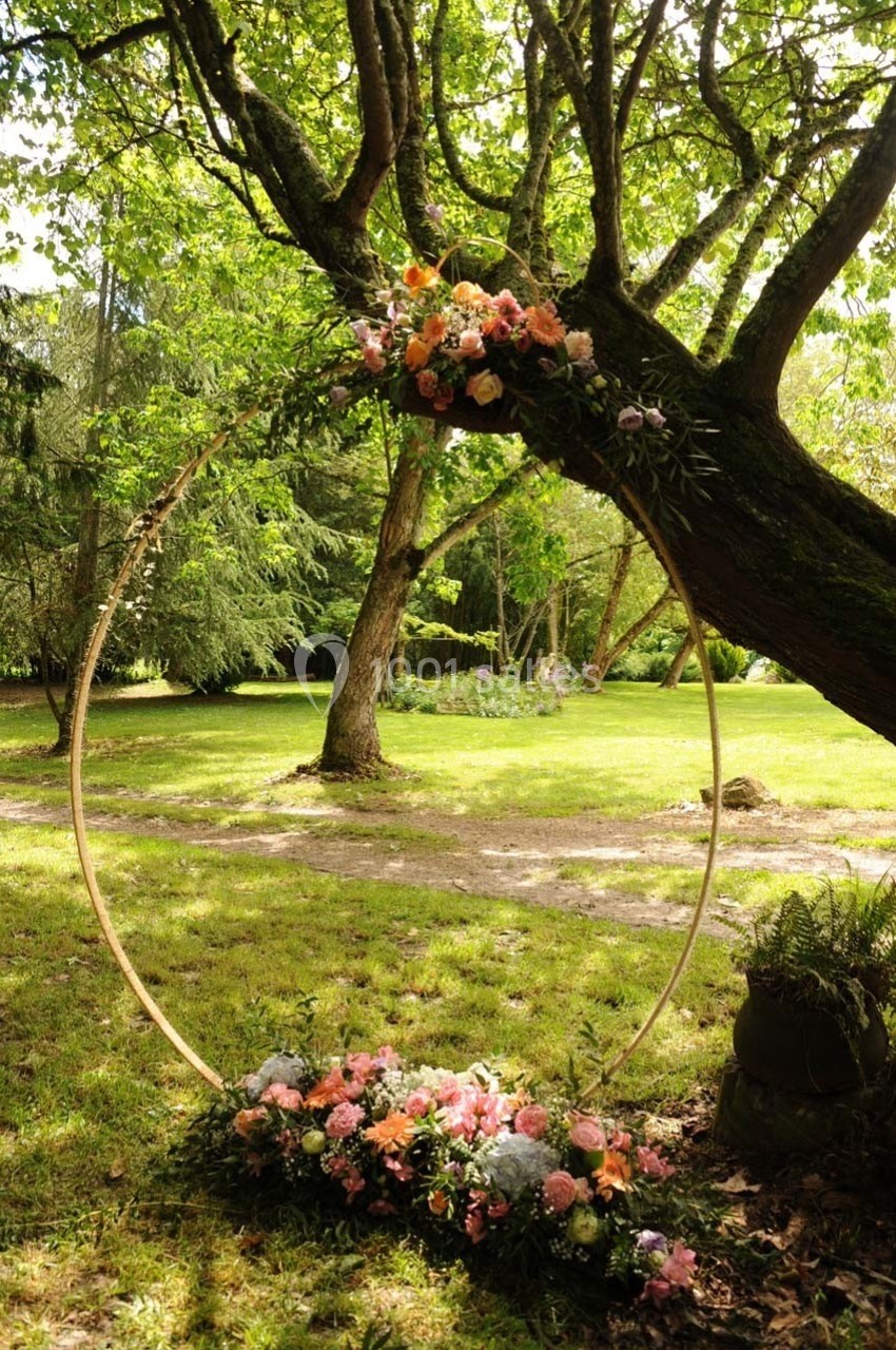 Cercle décoratif en bois orné de fleurs colorées, adossé à un arbre dans un jardin verdoyant.