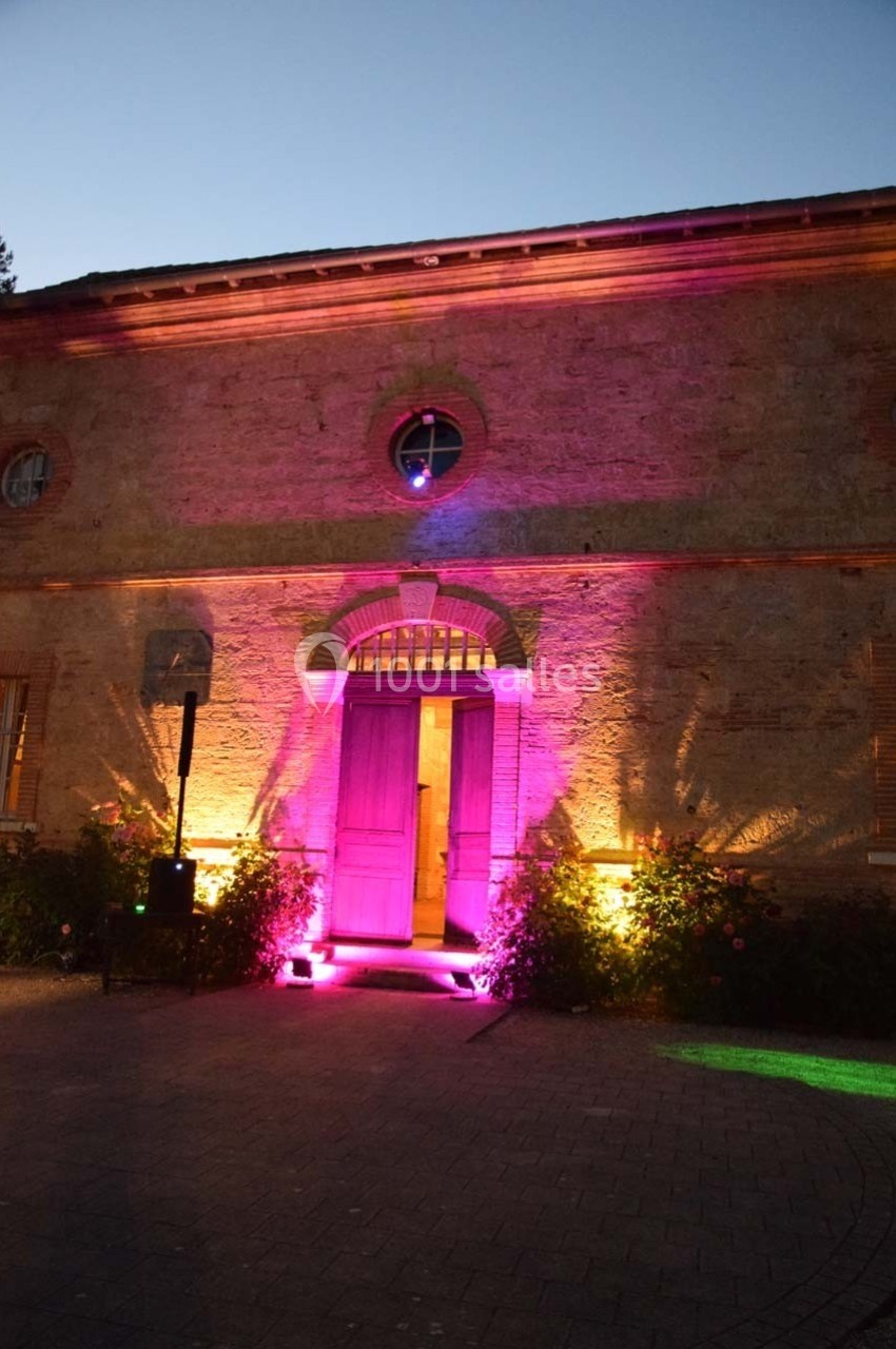 Façade en pierre d'un bâtiment éclairée par des lumières roses et jaunes au crépuscule.