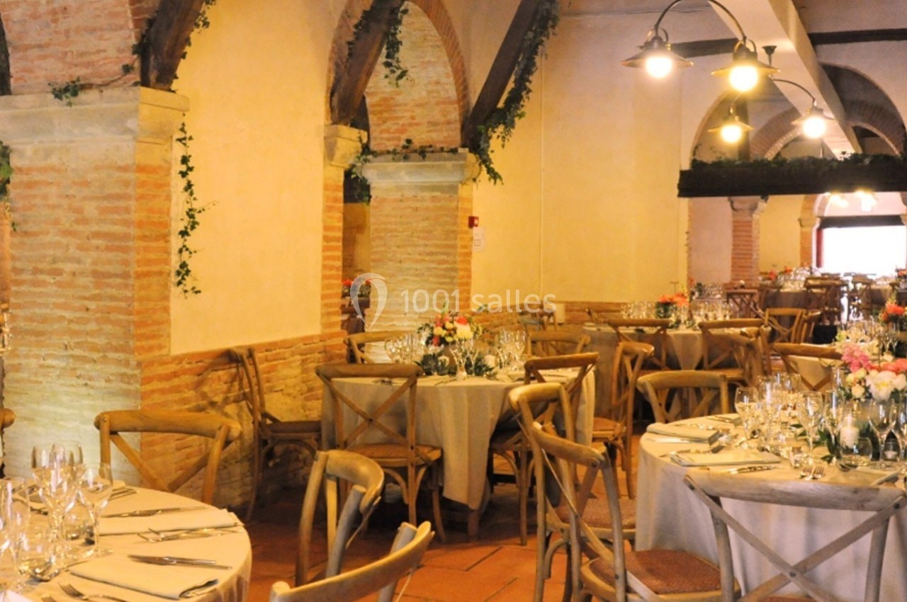 Salle de réception avec tables rondes dressées, chaises en bois, arches en briques et décoration florale.