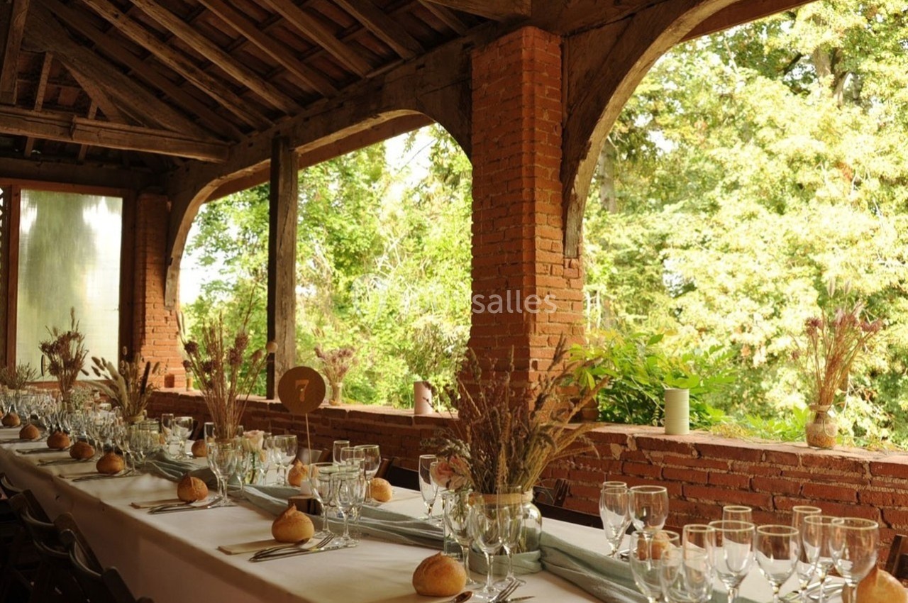 Grande table dressée avec vaisselle élégante, pains individuels et décoration florale, sous une terrasse en bois ouverte sur…